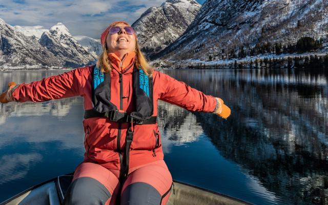 A woman in a red jacket sits on the boat with her arms outstretched and her face turned to the sun, the calm fjord mirroring the snow-covered mountains.