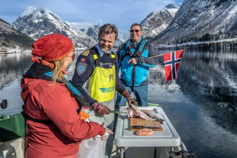 Three people in the winter sun on the boat, one filming with a phone, a guide in a yellow Varde vest pointing and a guest holding the Norwegian flag, with a filleted fish and a crab on the table and snowy mountains mirrored in the fjord.