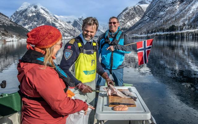 Three people in the winter sun on the boat, one filming with a phone, a guide in a yellow Varde vest pointing and a guest holding the Norwegian flag, with a filleted fish and a crab on the table and snowy mountains mirrored in the fjord.