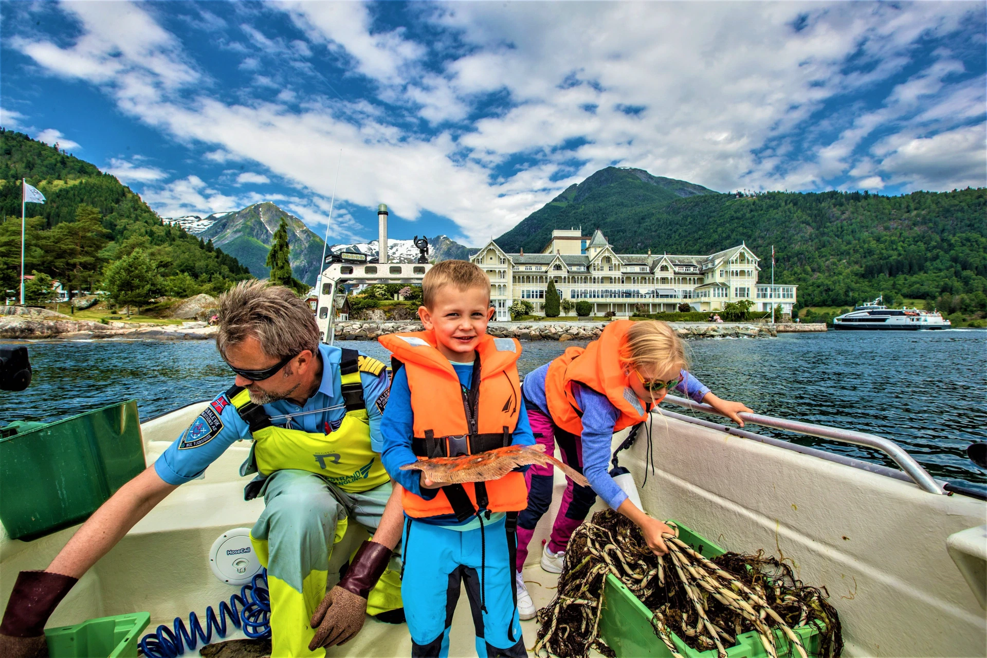 On the boat in front of the white hotel on the Balestrand shore the skipper lifts a green fish box while a boy in an orange life vest holds up a small flatfish and a girl reaches into a box of kelp.