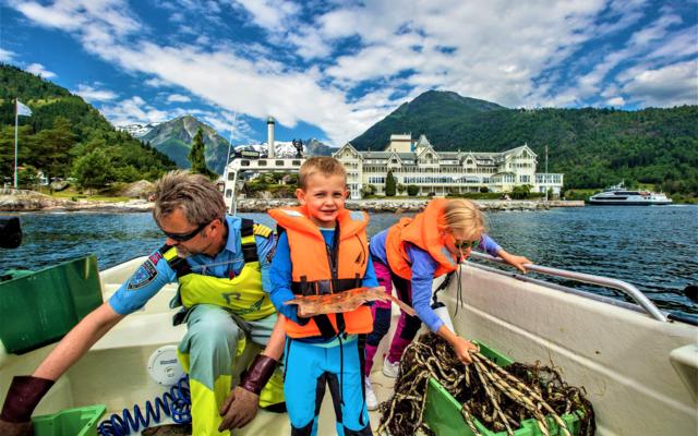 On the boat in front of the white hotel on the Balestrand shore the skipper lifts a green fish box while a boy in an orange life vest holds up a small flatfish and a girl reaches into a box of kelp.