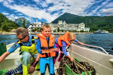 On the boat in front of the white hotel on the Balestrand shore the skipper lifts a green fish box while a boy in an orange life vest holds up a small flatfish and a girl reaches into a box of kelp.