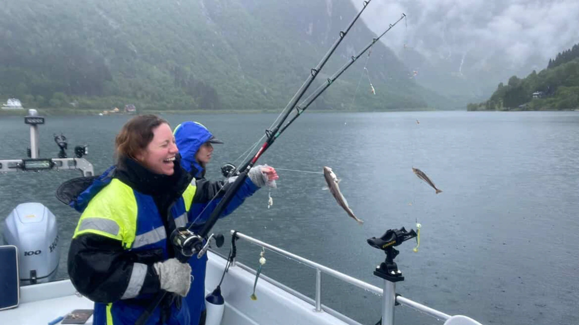 "Two Italian women from New York City experiencing the raw beauty of a rainy day on the Sognefjord with Balestrand Fjord Angling. One woman is laughing with pure joy while reeling in a fresh catch (whiting) on a fishing rod. Authentic Norwegian nature experience with misty mountains and rain, showcasing that 'Simplicity Creates Emotions' regardless of the weather. Highlighting adventure travel, female empowerment in the outdoors, and the 'Human Edge' in Balestrand, Norway."