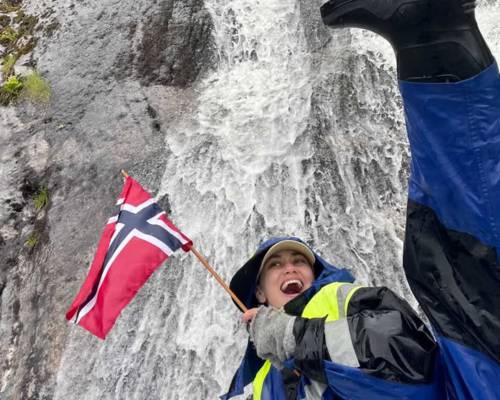 "A happy female guest smiling on a guided boat trip with Balestrand Fjord Angling in the Sognefjord, Norway. She is holding a Norwegian flag, wearing professional flotation gear and boots, with a powerful waterfall in the background. The scene showcases the pure joy of 'Simplicity Creates Emotions' and authentic 'Human Edge' experiences in Balestrand, Norway."