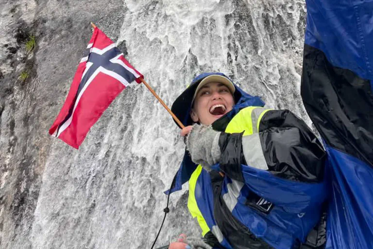 "A happy female guest smiling on a guided boat trip with Balestrand Fjord Angling in the Sognefjord, Norway. She is holding a Norwegian flag, wearing professional flotation gear and boots, with a powerful waterfall in the background. The scene showcases the pure joy of 'Simplicity Creates Emotions' and authentic 'Human Edge' experiences in Balestrand, Norway."