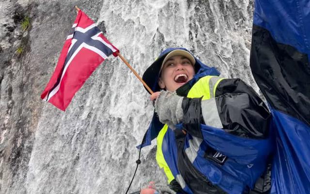 "A happy female guest smiling on a guided boat trip with Balestrand Fjord Angling in the Sognefjord, Norway. She is holding a Norwegian flag, wearing professional flotation gear and boots, with a powerful waterfall in the background. The scene showcases the pure joy of 'Simplicity Creates Emotions' and authentic 'Human Edge' experiences in Balestrand, Norway."