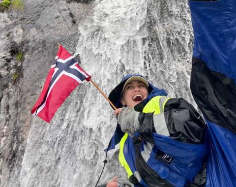 "A happy female guest smiling on a guided boat trip with Balestrand Fjord Angling in the Sognefjord, Norway. She is holding a Norwegian flag, wearing professional flotation gear and boots, with a powerful waterfall in the background. The scene showcases the pure joy of 'Simplicity Creates Emotions' and authentic 'Human Edge' experiences in Balestrand, Norway."