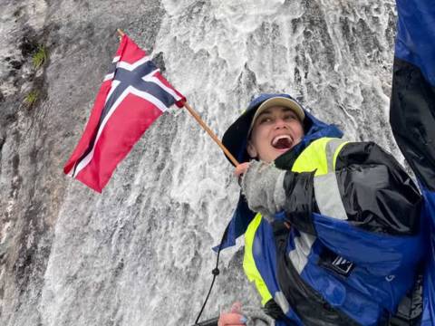"A happy female guest smiling on a guided boat trip with Balestrand Fjord Angling in the Sognefjord, Norway. She is holding a Norwegian flag, wearing professional flotation gear and boots, with a powerful waterfall in the background. The scene showcases the pure joy of 'Simplicity Creates Emotions' and authentic 'Human Edge' experiences in Balestrand, Norway."