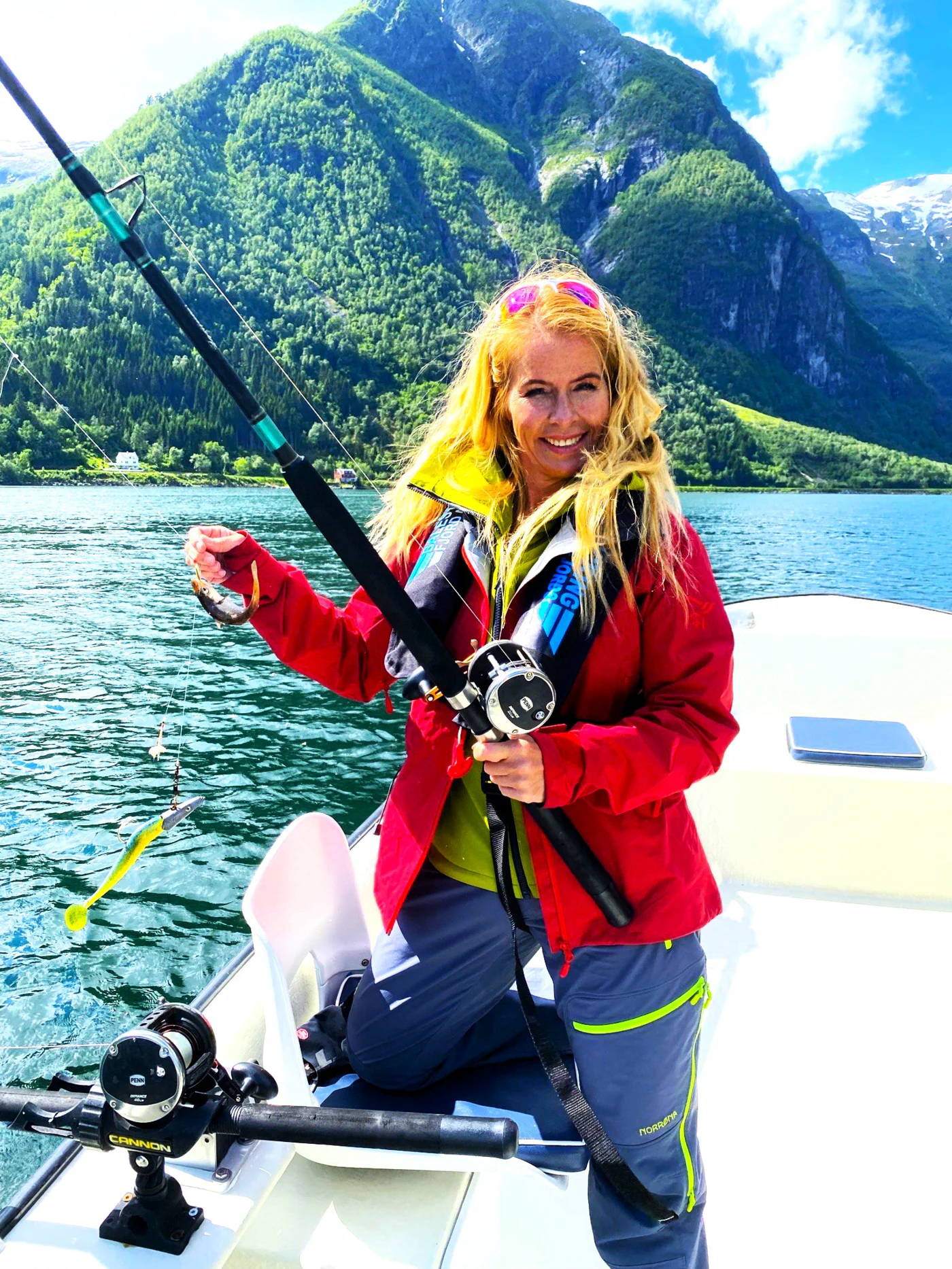 The fjord chicken challenge in Esefjord og Sognefjord. Blond dame i rød jakke, smiler blant høye fjell og solskinn og vind i håret