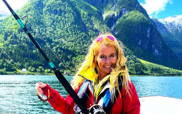 A smiling woman in a red jacket holds a fishing rod on the boat, the fjord and steep green mountains behind.