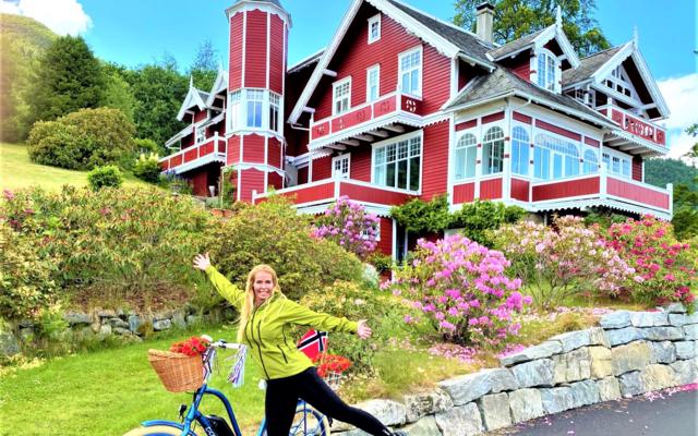 A woman with an electric bike smiling in front of a historic red dragon-style villa in Balestrand, showcasing family-friendly bike rentals and shore excursions near Vik cruise port.