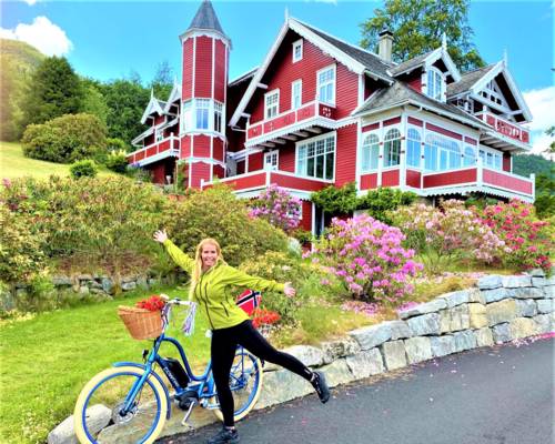 A woman with an electric bike smiling in front of a historic red dragon-style villa in Balestrand, showcasing family-friendly bike rentals and shore excursions near Vik cruise port.
