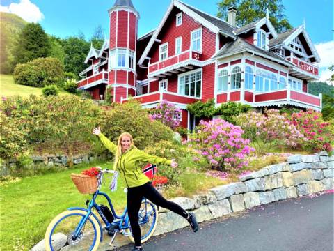 A woman with an electric bike smiling in front of a historic red dragon-style villa in Balestrand, showcasing family-friendly bike rentals and shore excursions near Vik cruise port.