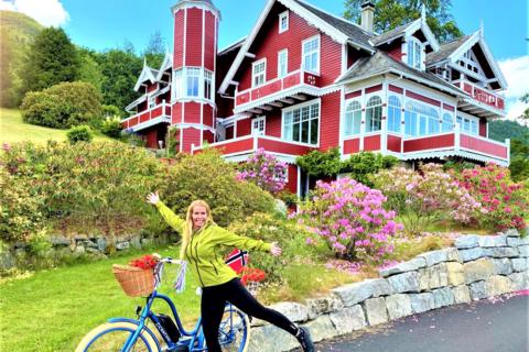 A woman with an electric bike smiling in front of a historic red dragon-style villa in Balestrand, showcasing family-friendly bike rentals and shore excursions near Vik cruise port.