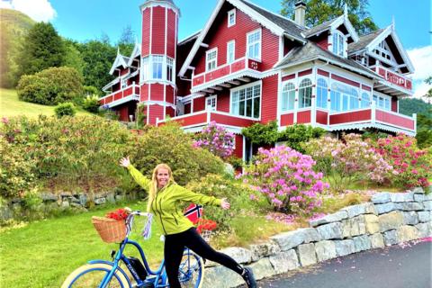 A woman with an electric bike smiling in front of a historic red dragon-style villa in Balestrand, showcasing family-friendly bike rentals and shore excursions near Vik cruise port.