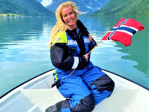 A smiling person with blonde hair kneels on a white boat, holding a Norwegian flag. They are wearing a blue and black floatation suit. The boat is in the crisp, green water of a narrow fjord, surrounded by steep mountains under a cloudy sky. The scene is in Balestrand, Norway.