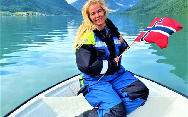 A smiling person with blonde hair kneels on a white boat, holding a Norwegian flag. They are wearing a blue and black floatation suit. The boat is in the crisp, green water of a narrow fjord, surrounded by steep mountains under a cloudy sky. The scene is in Balestrand, Norway.