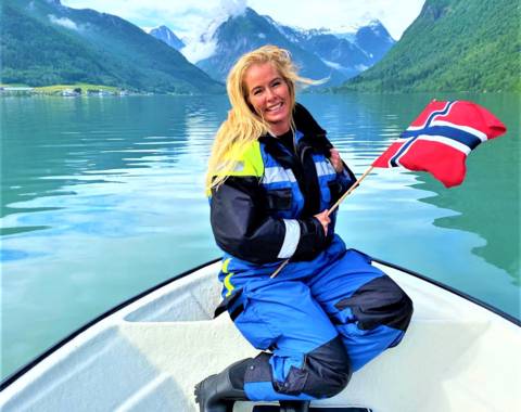 A smiling person with blonde hair kneels on a white boat, holding a Norwegian flag. They are wearing a blue and black floatation suit. The boat is in the crisp, green water of a narrow fjord, surrounded by steep mountains under a cloudy sky. The scene is in Balestrand, Norway.