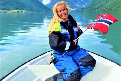 A smiling person with blonde hair kneels on a white boat, holding a Norwegian flag. They are wearing a blue and black floatation suit. The boat is in the crisp, green water of a narrow fjord, surrounded by steep mountains under a cloudy sky. The scene is in Balestrand, Norway.