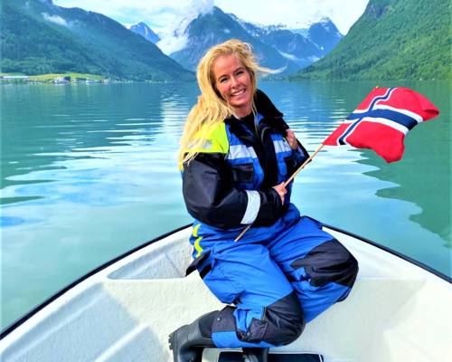 A smiling person with blonde hair kneels on a white boat, holding a Norwegian flag. They are wearing a blue and black floatation suit. The boat is in the crisp, green water of a narrow fjord, surrounded by steep mountains under a cloudy sky. The scene is in Balestrand, Norway.