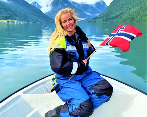 A smiling person with blonde hair kneels on a white boat, holding a Norwegian flag. They are wearing a blue and black floatation suit. The boat is in the crisp, green water of a narrow fjord, surrounded by steep mountains under a cloudy sky. The scene is in Balestrand, Norway.