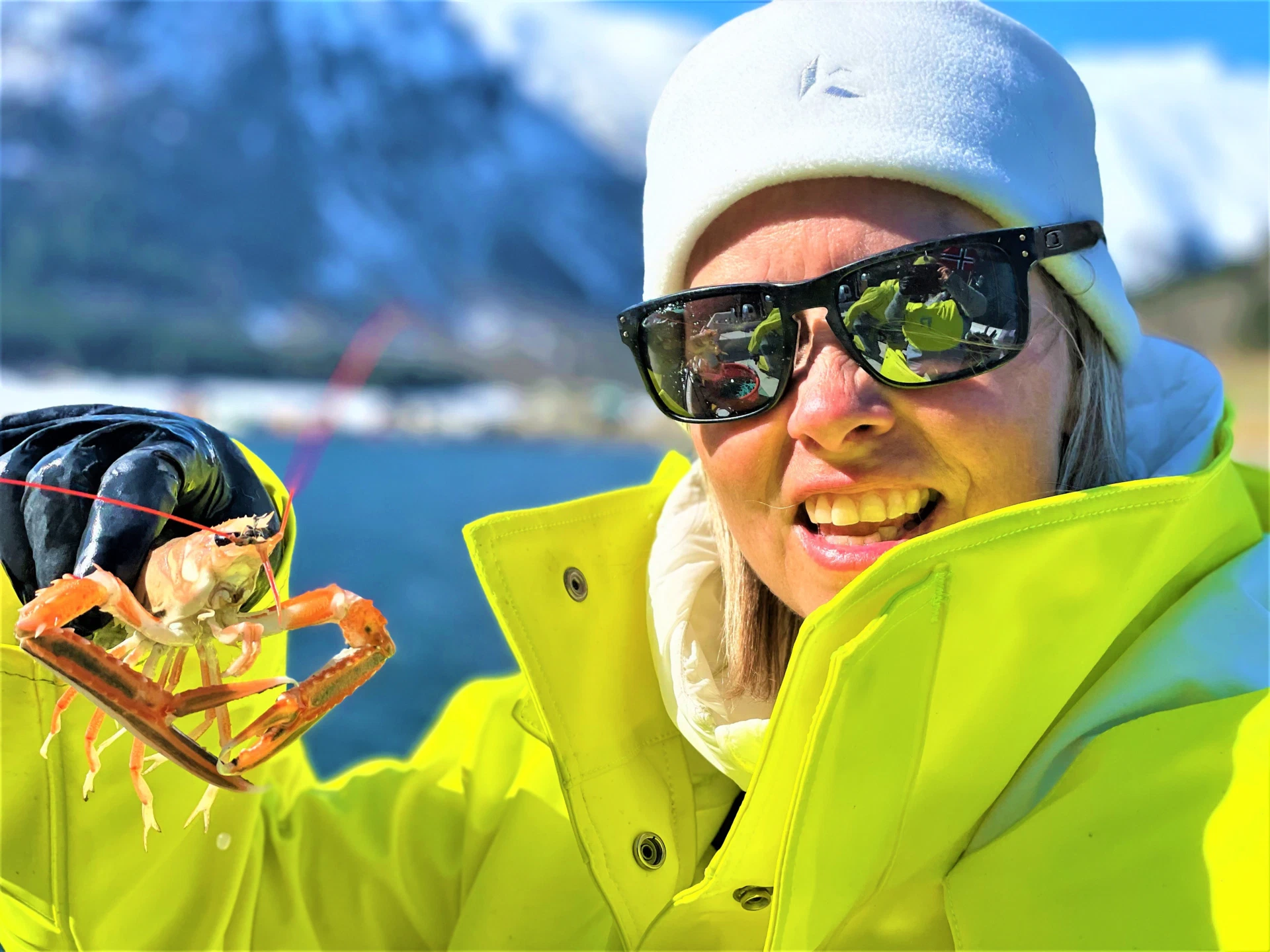 A woman in a yellow winter jacket and white beanie holds a live langoustine in her gloved hand, snowy mountains and blue fjord water behind.