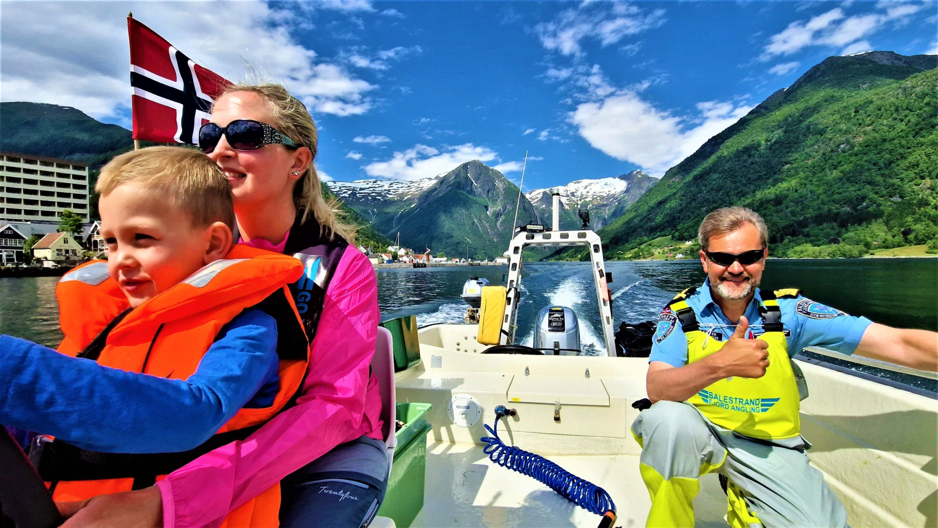 A boy in an orange life vest looks at the camera while the skipper gives a thumbs up at the wheel, the Norwegian flag flying and the village of Balestrand on the shore behind.