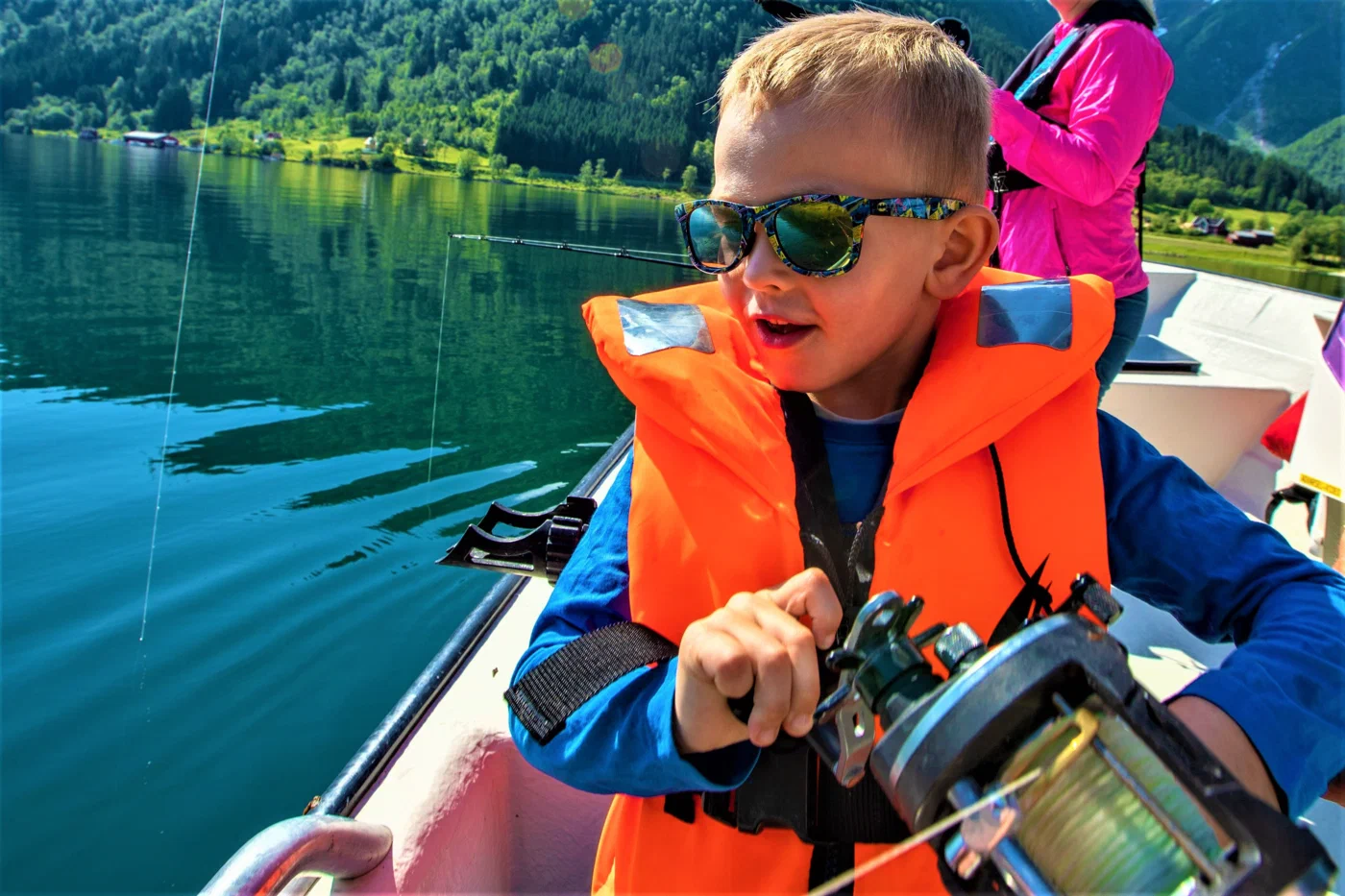 A boy in an orange life vest and sunglasses reels in a fishing rod on the boat, the line running out over the fjord water.