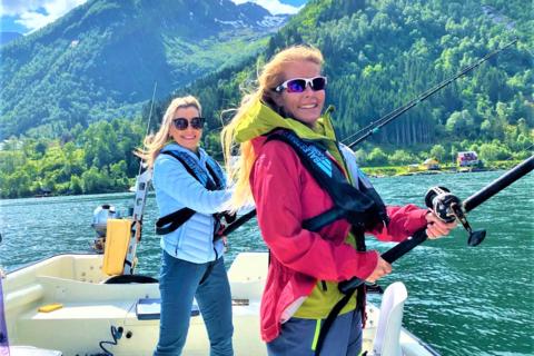Two smiling women in life vests are fishing from a boat on a sunny day in a Norwegian fjord with steep green mountains in the background.