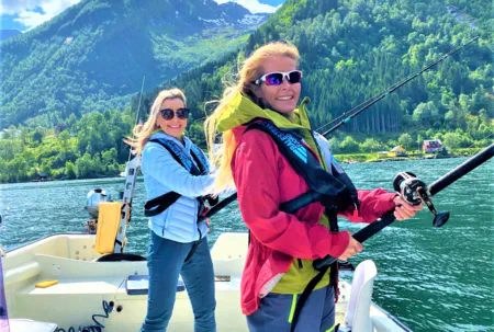 Two smiling women in life vests are fishing from a boat on a sunny day in a Norwegian fjord with steep green mountains in the background.