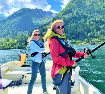 Two smiling women in life vests are fishing from a boat on a sunny day in a Norwegian fjord with steep green mountains in the background.