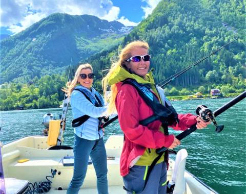 Two smiling women in life vests are fishing from a boat on a sunny day in a Norwegian fjord with steep green mountains in the background.