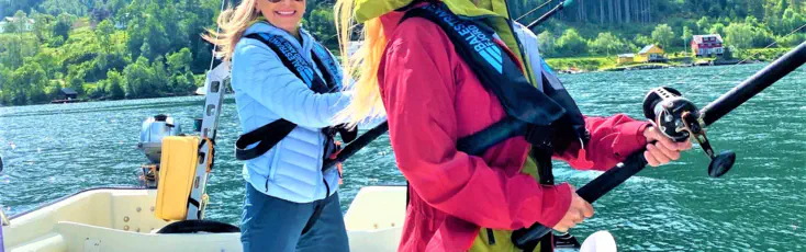 Two smiling women in life vests are fishing from a boat on a sunny day in a Norwegian fjord with steep green mountains in the background.