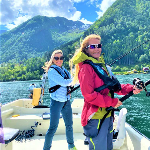 Two smiling women in life vests are fishing from a boat on a sunny day in a Norwegian fjord with steep green mountains in the background.