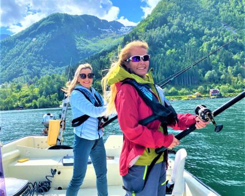 Two smiling women in life vests are fishing from a boat on a sunny day in a Norwegian fjord with steep green mountains in the background.