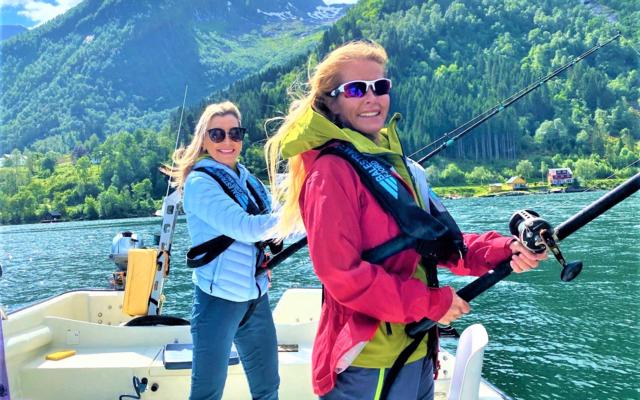 Two smiling women in life vests are fishing from a boat on a sunny day in a Norwegian fjord with steep green mountains in the background.